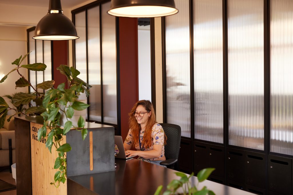 A woman in glasses sat at a metal reception desk with plants.