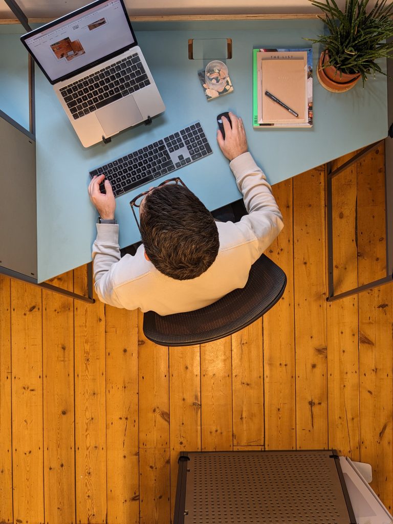 A birds eye view of a man sat at a desk in an office in front of a laptop.