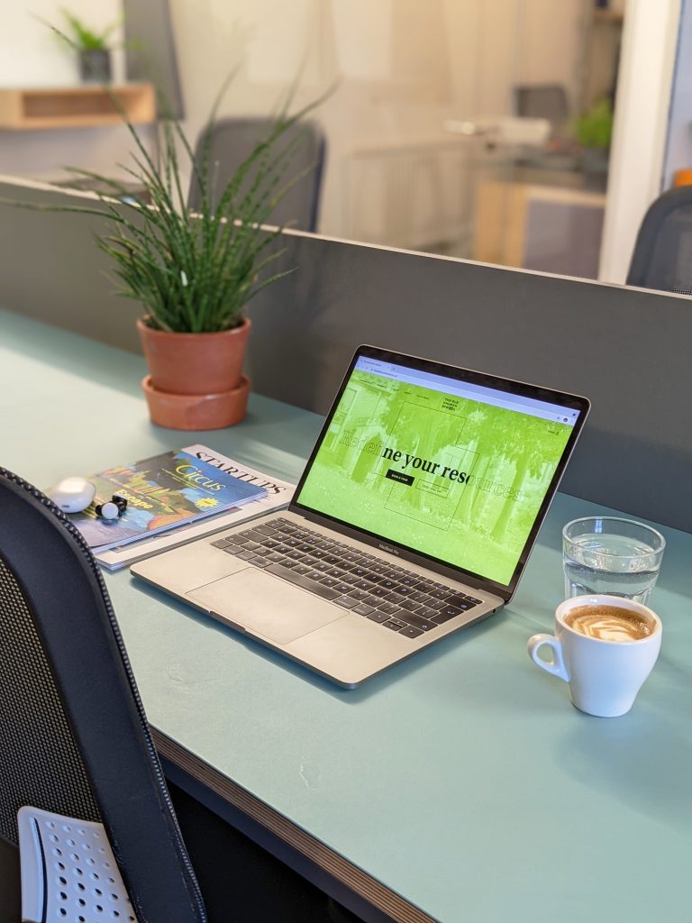 Close up of a laptop on a desk with a coffee, pot plant and magazine.