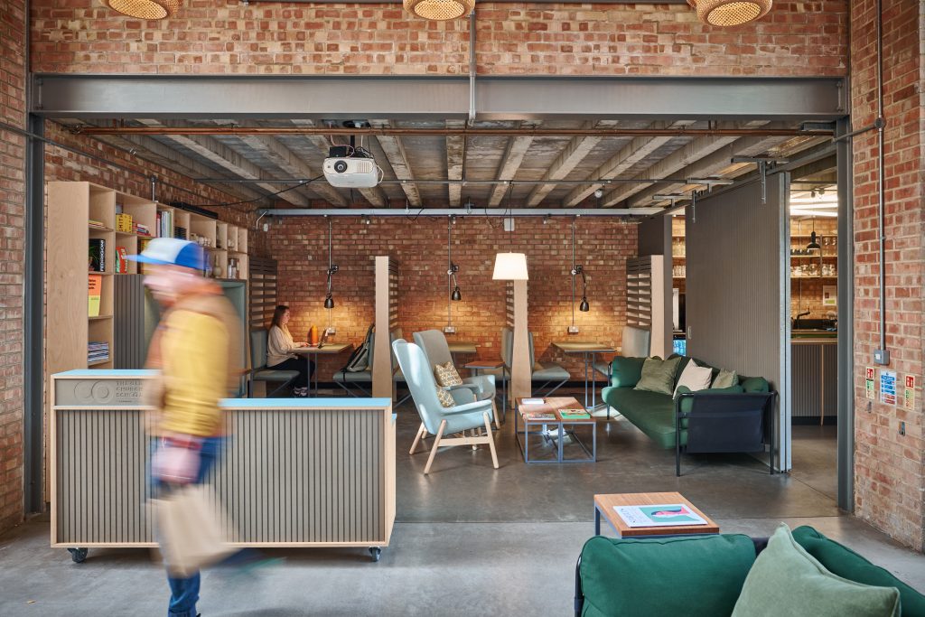 Open workspace with a green sofa, lady sat on her laptop and a man walking past