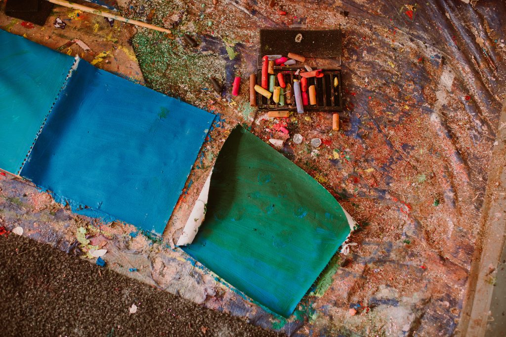 A messy floor of an artists studio with paint, oil pastels and paper.